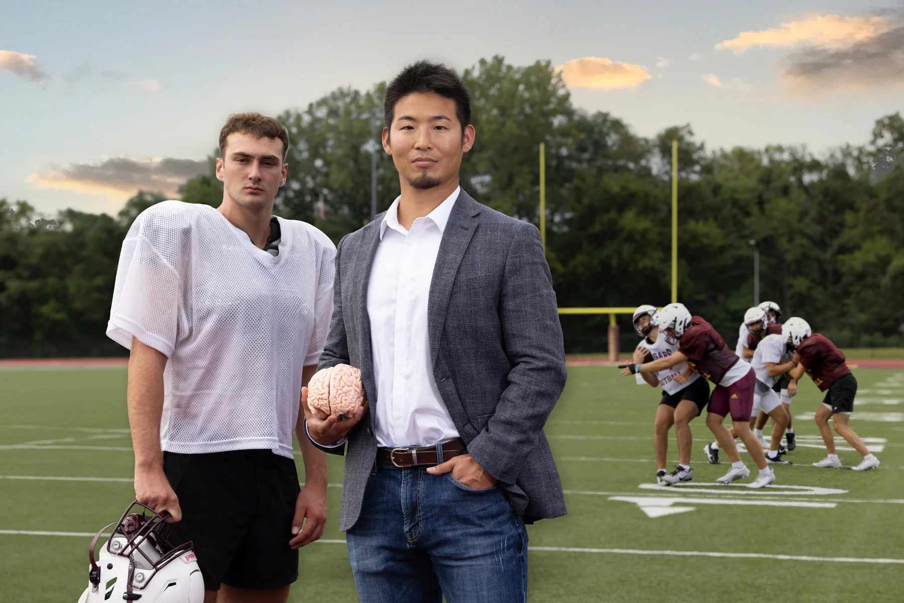 Kei Kawata stands with a high school player on a football field holding a brain while a scrimmage takes place in the background.